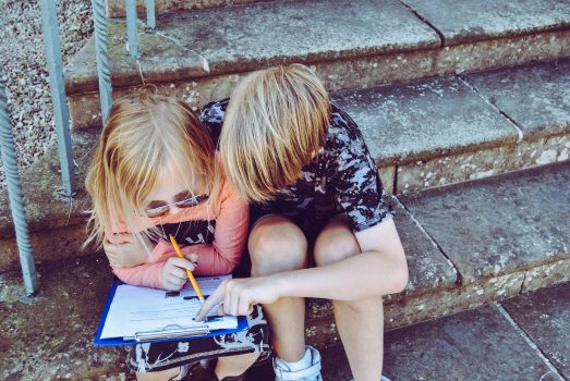 Na zdjęciu widać dwoje dzieci, chłopca i dziewczynkę. Siedzą na kamiennych schodach. Chłopiec pomaga dziewczynce w odrabianiu zadania domowego. 